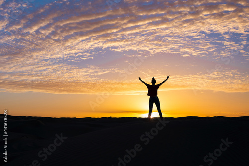Silhouette of a confident young woman standing in the sunset, arms raised. Power pose.