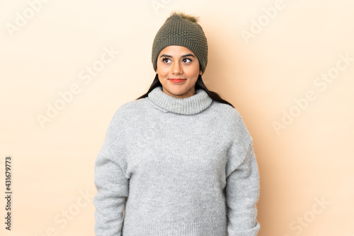 Young Colombian girl with winter hat isolated on beige background standing and looking to the side © luismolinero