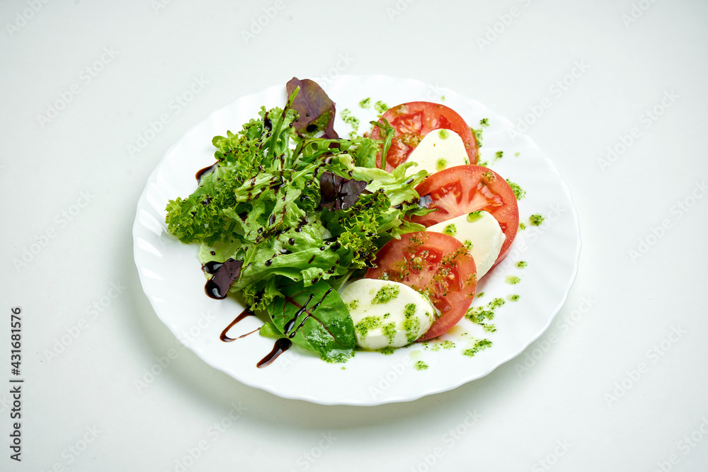 Classic Italian caprese salad with tomatoes, mozzarella, mix salad and pesto sauce in a white plate on a white plate