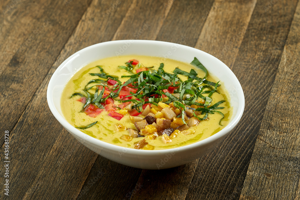 Appetizing and dietary dish - corn soup with cream and fried mushrooms, tomatoes and herbs in a white bowl on a wooden background Appetizing and dietary dish - corn soup with cream and fried mushrooms, tomatoes and herbs in a white bowl on a wooden background