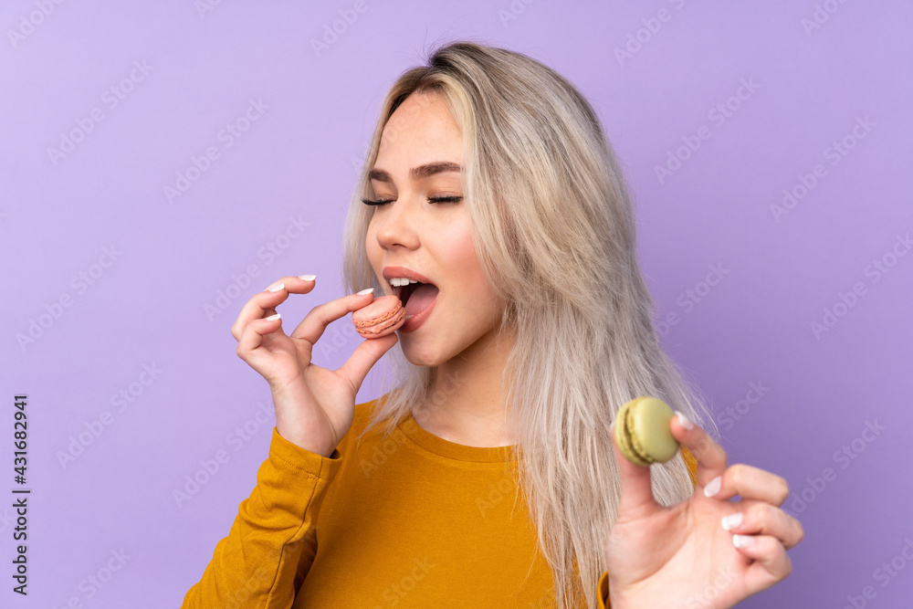 Teenager girl over isolated purple background holding colorful French macarons and eating it Teenager girl over isolated purple background holding colorful French macarons and eating it