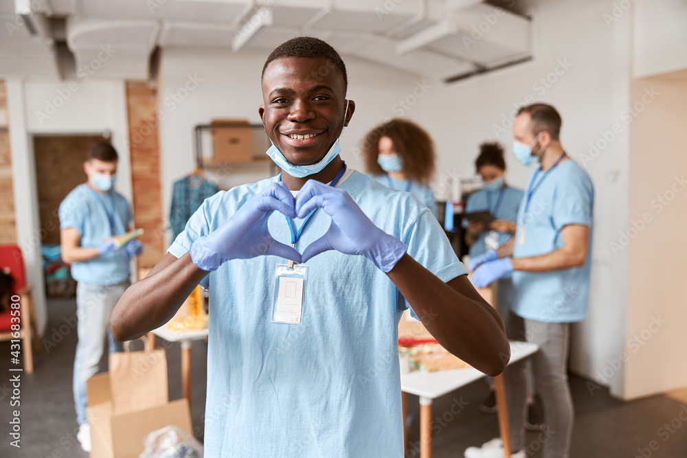 Obraz premium African american young male volunteer in blue uniform, protective mask and gloves smiling at camera and showing heart love sign. Team sorting, packing food stuff in the background
