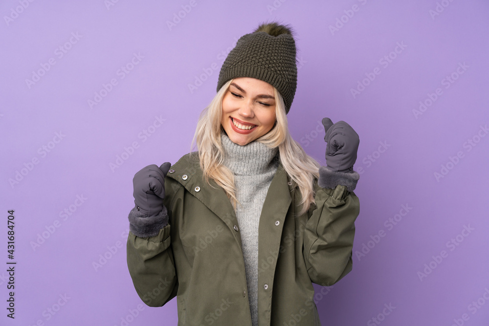 Teenager blonde girl with winter hat over isolated purple background celebrating a victory
