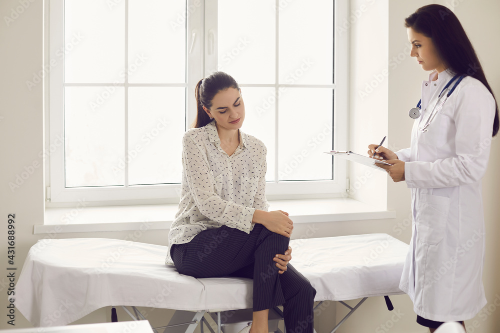 Patient suffering from knee pain visits doctor at clinic. Young woman sitting on physician's examination couch at hospital and complaining about painful inflammation in kneecap she injured in accident Patient suffering from knee pain visits doctor at clinic. Young woman sitting on physician's examination couch at hospital and complaining about painful inflammation in kneecap she injured in accident