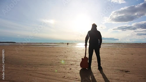 A man with a guitar stands with his back on the beach and looks at the sea.
