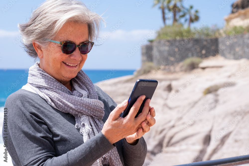 Attractive senior woman white-haired using smartphone outdoors at the sea smiling. Joyful lifestyles concept, happy retirement