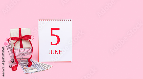 A gift box in a shopping trolley, dollars and a calendar with the date of 5 june on a pink background. photo