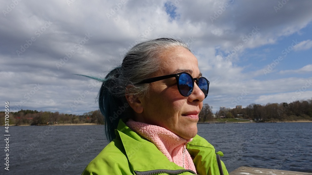 Photo of a woman's face with ponytail hair, sunglasses, a pink sweatshirt and a light green jacket