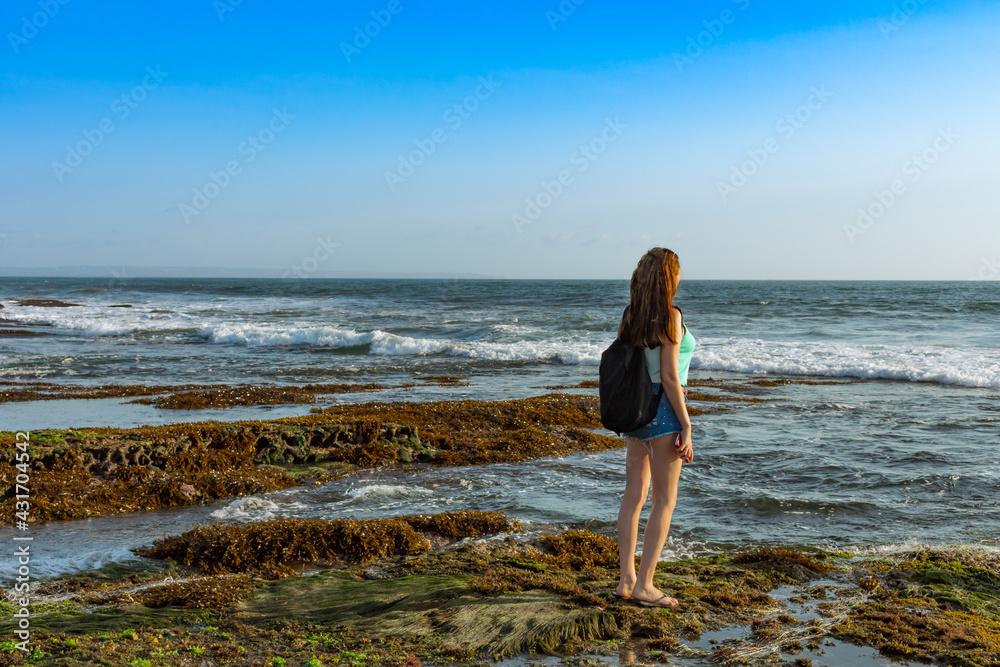 A slender young traveler girl with a black backpack on her shoulders looks out to sea towards the sunset near the Tanah Lot Temple in Bali, Indonesia. A woman in short denim shorts and a green tank