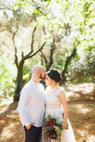 The bride and groom with a bouquet stand hugging among the trees in an olive grove, the groom kisses the bride on the forehead 