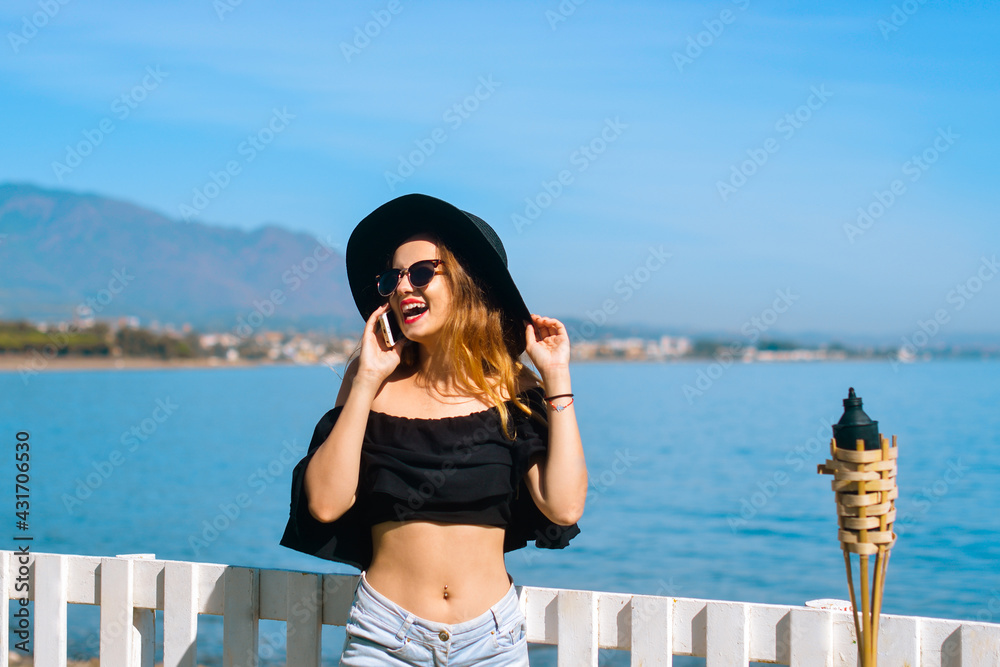 A beautiful girl speaks on the phone against the backdrop of the beach and ocean, sea. Woman in a big black hat and glasses on vacation. Freelancer calls and smiles, roaming and chatting. Travel