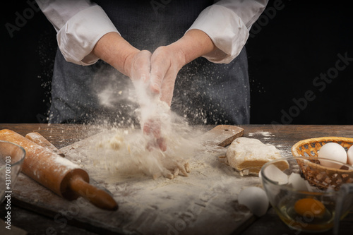 Women's hands, flour and dough. A woman in an apron cooking dough for homemade baking, a rustic home cozy atmosphere, a dark background with unusual lighting.