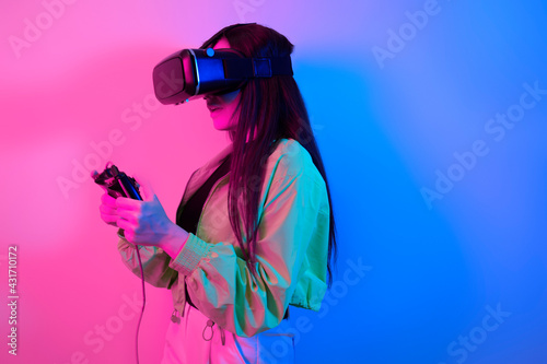 Laughing smiling surprise girl in neon with VR glasses holds a gamepad in her hands and plays games. Virtual world, technology.