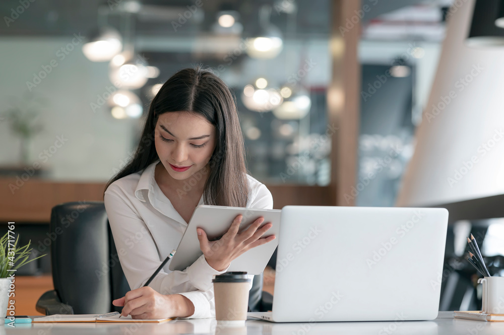 Young beautiful asian woman working on tablet and laptop computer while sitting at her desk office.