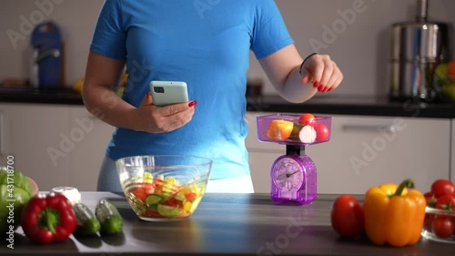 Midsection of healthy eating woman using kitchen scales to weigh vegetables and smartphone with weight loss application to calculate calorie content of dish. Slimming woman counting calories