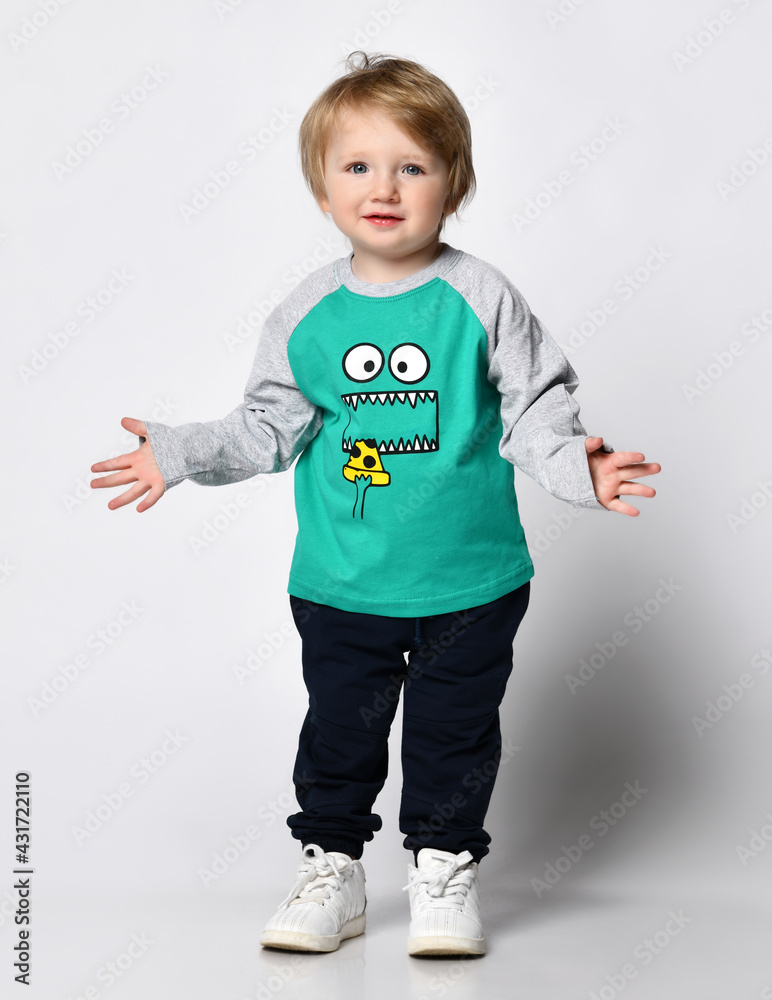 small smiling preschooler of European descent in a trendy green raglan with cute black sweatpants with a print on the face stands and spreads his arms. Studio portrait on white small smiling preschooler of European descent in a trendy green raglan with cute black sweatpants with a print on the face stands and spreads his arms. Studio portrait on white