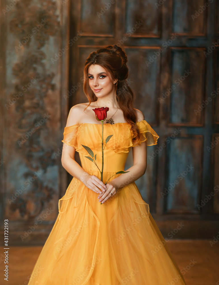 Girl beauty fantasy princess in yellow long historical, medieval silk dress  holding flower red rose in her hands. Background of old gothic castle room.  Fairy tale bewitched queen. Happy woman smiles Stock-Foto