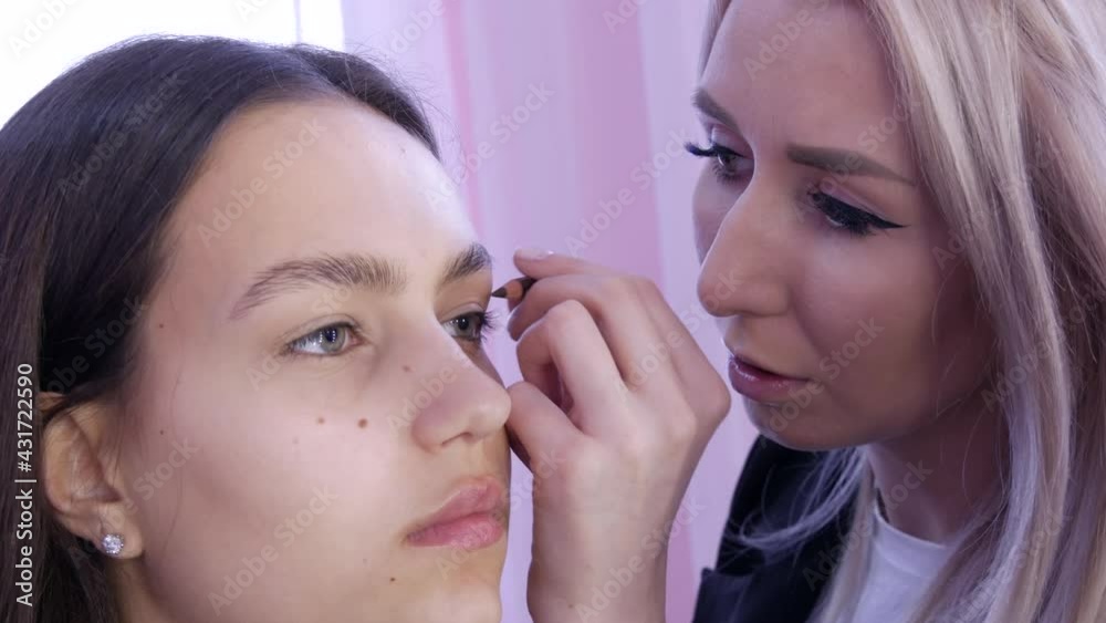 The stylist make-up master makes an eye on the evening stylish bright makeup on a young girl model in a beauty studio, a special brush paints with eye shadows using a special pencil The stylist make-up master makes an eye on the evening stylish bright makeup on a young girl model in a beauty studio, a special brush paints with eye shadows using a special pencil