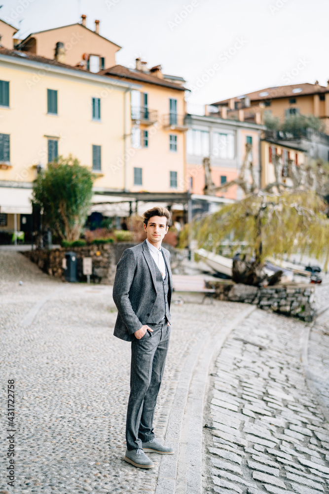 Groom stands with his hands in his pockets on a cobbled street against the backdrop of ancient villas. Lake Como