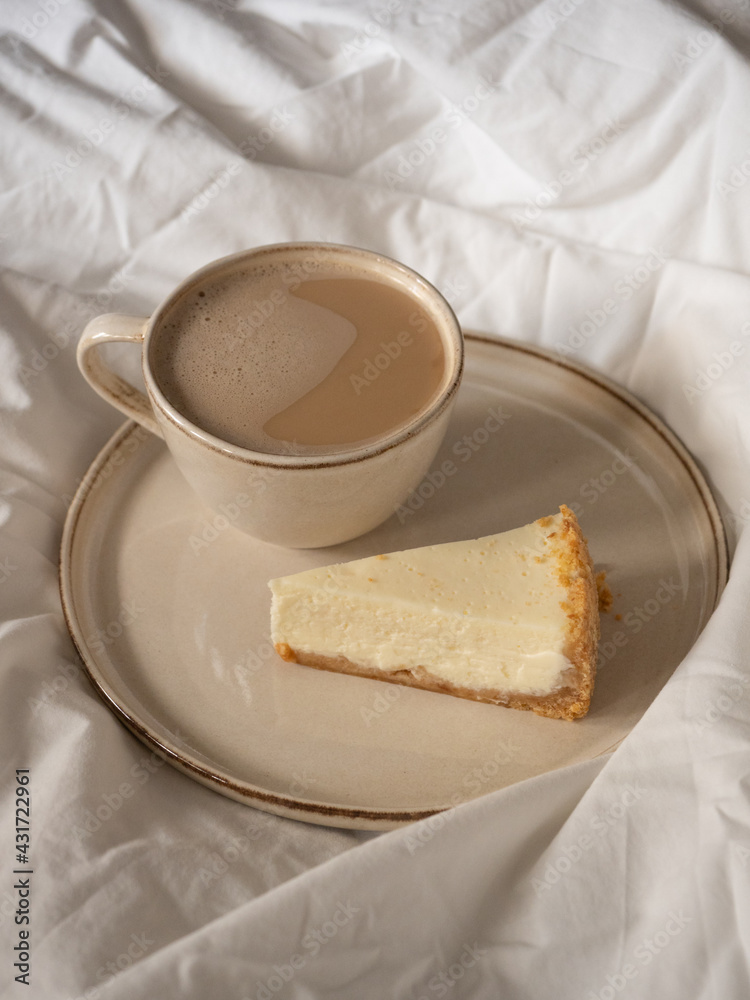 Morning cup of coffee and classic cheesecake on a beige ceramic plate on a soft white bed in the morning light Morning cup of coffee and classic cheesecake on a beige ceramic plate on a soft white bed in the morning light