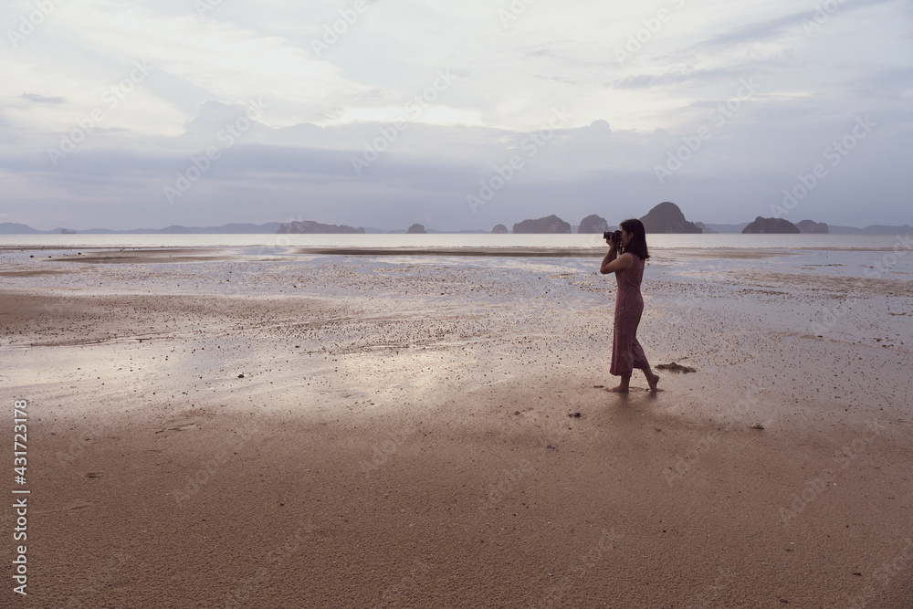 young asian woman using camera to shoot photo of sunset on beach in moody day