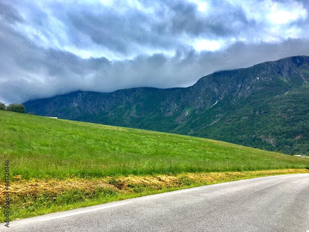 Blurry Cloudy Sky, Mountain Hill, Green Field And Asphalt Road In Triangular Proportions . Skjolden, Norway.  June 23, 2019