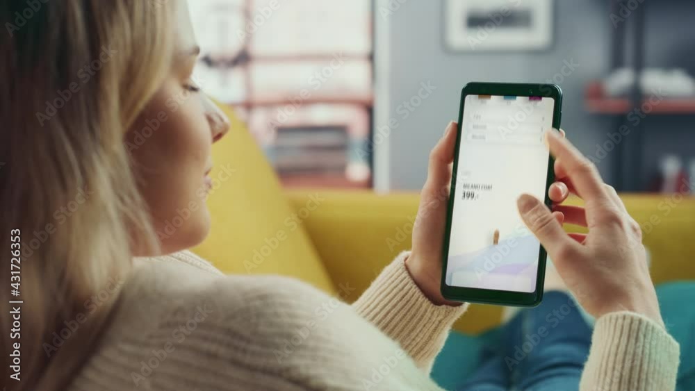 Beautiful Authentic Female Online Shopping on Her Smartphone at Home Living Room while Lying on a Couch Sofa. She's Browsing the Internet and Checking Deals in Her Favourite Internet Clothing Store.