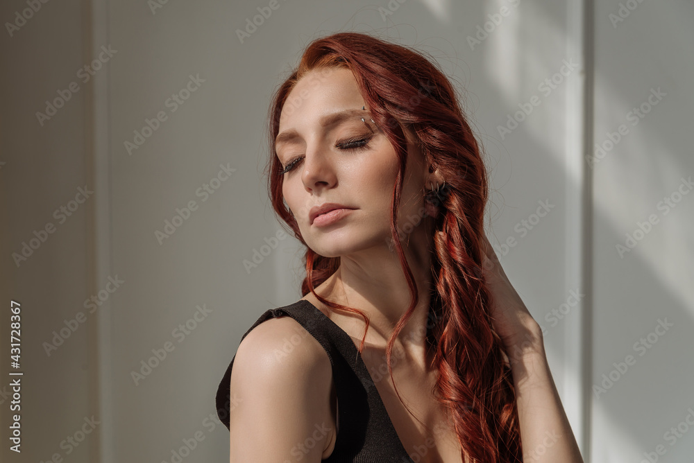 an attractive red-haired woman with her eyes closed enjoys the sunlight from the window. close-up portrait of a beautiful woman with her eyes closed. the concept of calm, harmony