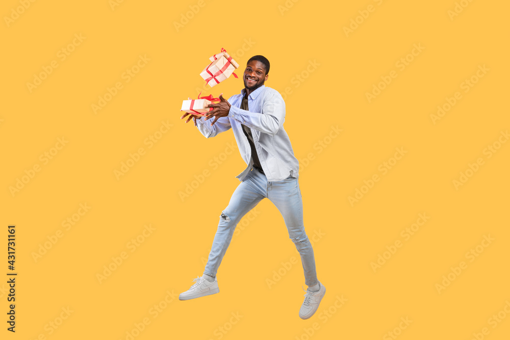 Holidays and celebration time. Overjoyed african american guy holding gift boxes and jumping on yellow background