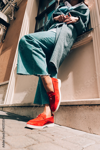 Woman in a red stylish suede loafer shoes, blue jeanse, white shirt and trendy checkered coat posing on the street. Pair of trendy female loafers shoes. photo