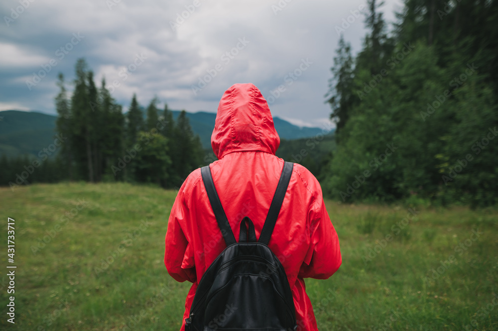 Person in the red raincoat and backpack stands in the mountains in the mountain valley, the view from the back. Girl tourist walks the mountains on a cloudy day. Person in the red raincoat and backpack stands in the mountains in the mountain valley, the view from the back. Girl tourist walks the mountains on a cloudy day.