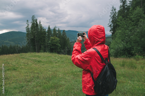 Hiker woman in red raincoat and with backpack makes photo of beautiful mountain scenery on smartphone. Tourist tourist takes photos of mountain landscapes and walks on mountain.
