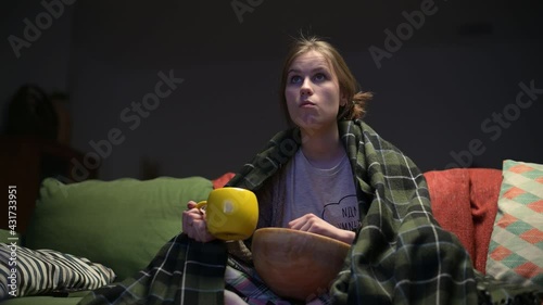 Lonely young woman watching TV in the evening, covered with a blanket and eating chips instead of going to bed