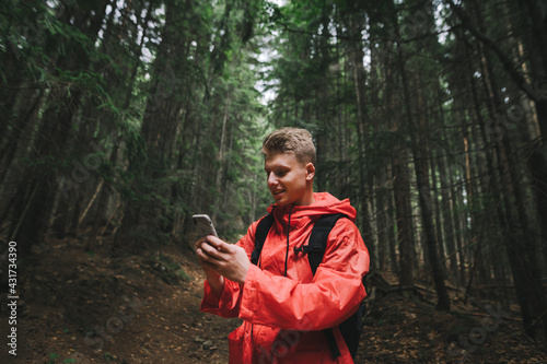Smiling young man in a red raincoat stands on a forest path in the mountains and uses a smartphone with a positive face. Happy mountain hiker uses smartphone, stands in dark coniferous forest.