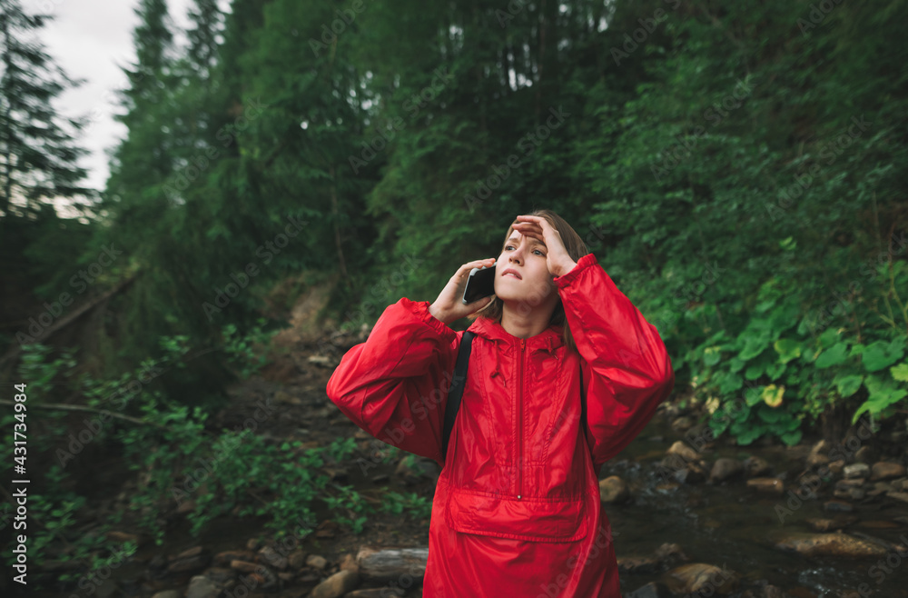 Concerned girl tourist stands in the forest in a red raincoat, calls on phone and looks away with a worried face in a mountain hike against a backdrop of a mountain stream and coniferous trees.