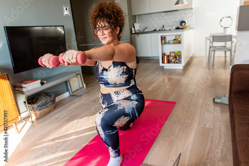 Young plus size woman at home doing exercise in front of open laptop, repeating instructions by professional online fitness trainer.
