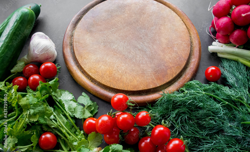 A pile of fresh, ripe vegetables: radishes, tomatoes, cucumbers, herbs, and garlic are arranged around a round wooden chopping board. Copy space A pile of fresh, ripe vegetables: radishes, tomatoes, cucumbers, herbs, and garlic are arranged around a round wooden chopping board. Copy space