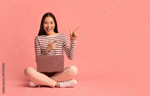 Obraz na plátně Asian woman sitting on floor with laptop and pointing at copy space