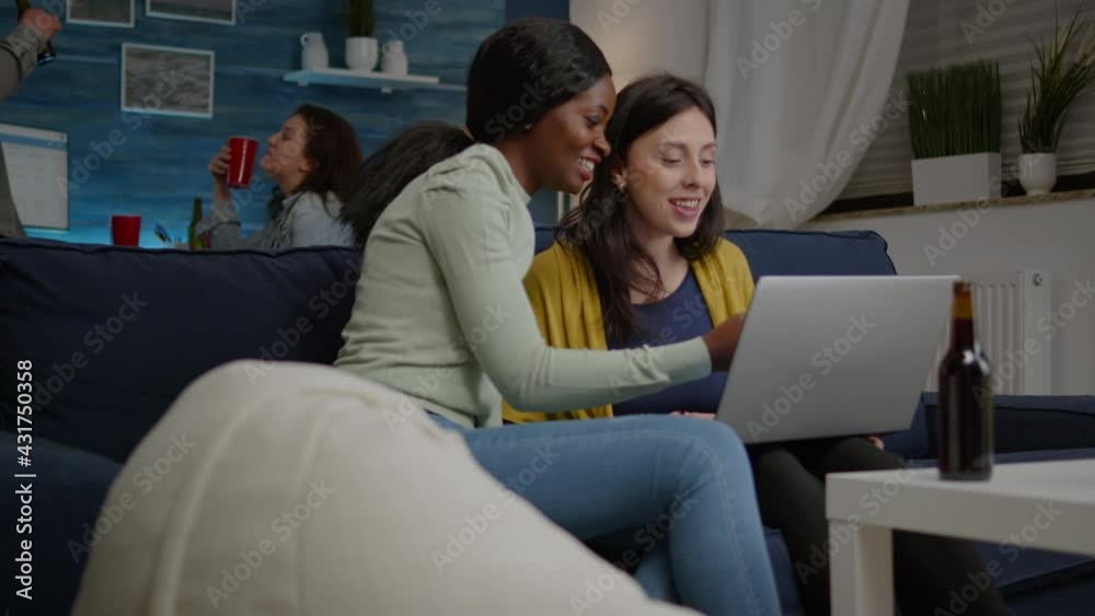 Mixed race friends talking while looking at funny video series on laptop sitting on couch in living room. In background woman and man drinking beer enjoying time together during fun party Mixed race friends talking while looking at funny video series on laptop sitting on couch in living room. In background woman and man drinking beer enjoying time together during fun party