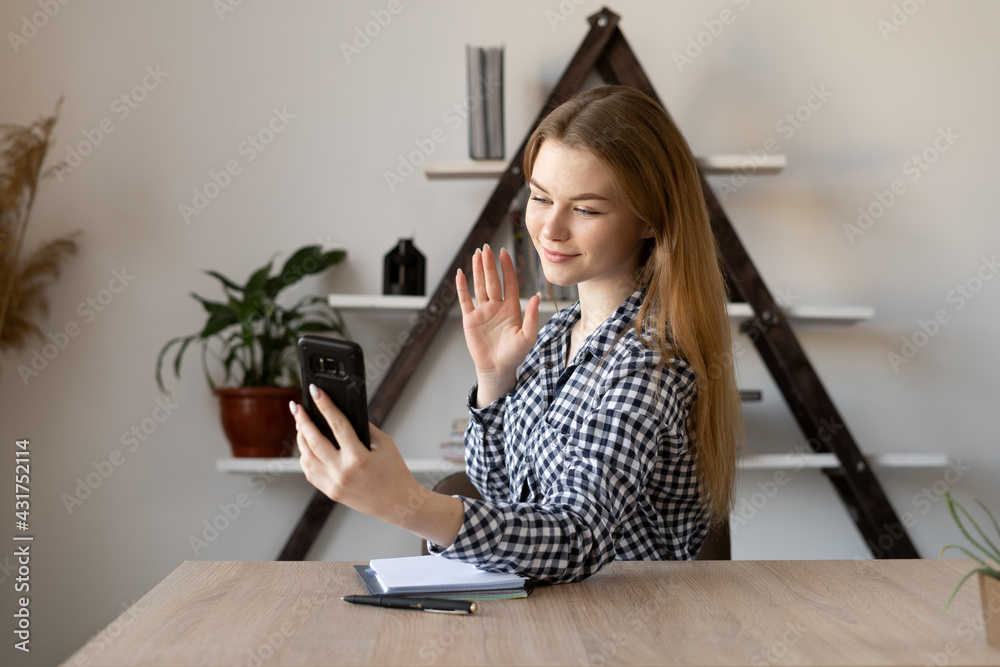 Smiling Caucasian young woman waves at smartphone camera hello and records video vlog about life. Millennial lady girl is sitting in the office and watching a video greeting. Smiling Caucasian young woman waves at smartphone camera hello and records video vlog about life. Millennial lady girl is sitting in the office and watching a video greeting.