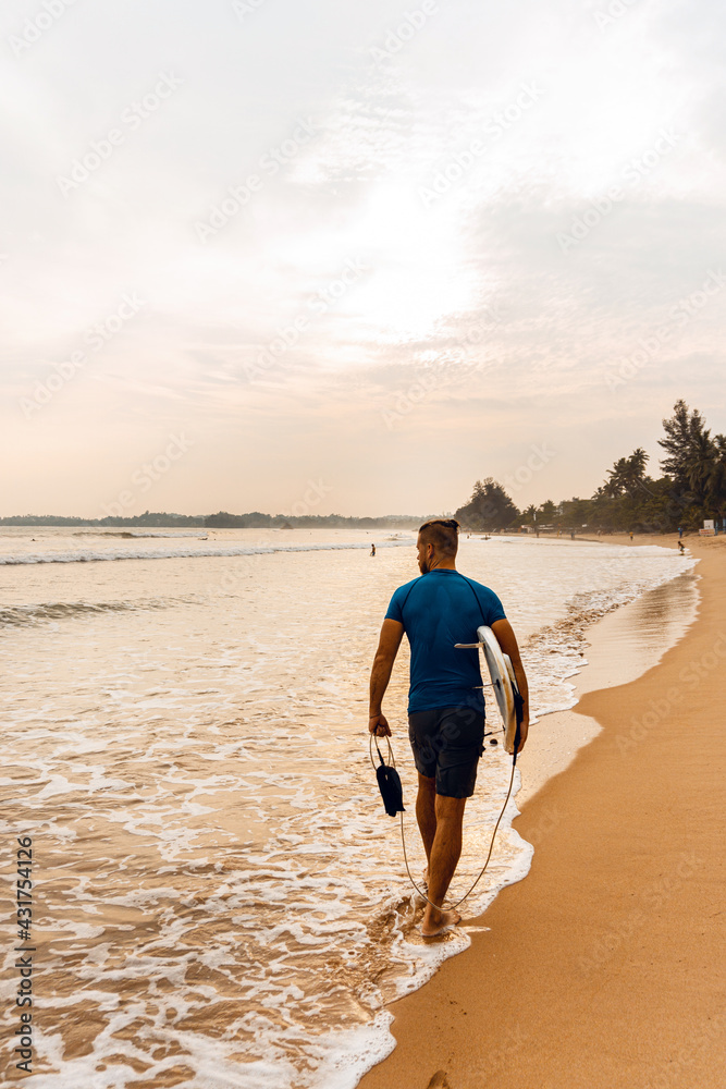 Surfer mężczyzna idący z deską po plaży na tle zachodu słońca. - obrazy, fototapety, plakaty 