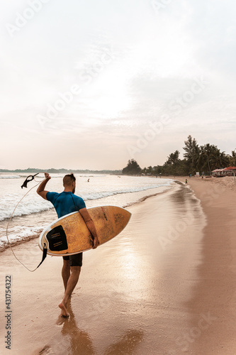 Surfer mężczyzna idący z deską po plaży na tle zachodu słońca.