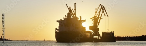 Large cargo ship (bulk carrier) anchored in port at sunset, close-up. Cranes in the background. Riga, Baltic sea, Latvia. Freight transportation, global communications, logistics, environmental damage photo