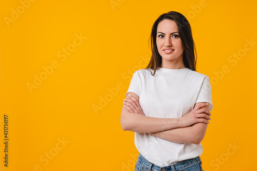 Smiling young caucasian woman in white T-shirt crossing her arms isolated on yellow background. Cheerful latin-american 30s woman standing with arms crossed