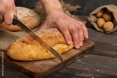 Male hands with a bread knife cut fresh crispy baguette on a wooden board. Sliced traditional french bread. Close-up