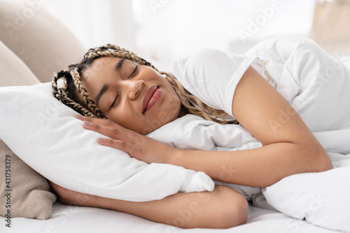 Fototapeta Cheerful young african woman lying and sleeping in her white bed in the morning
