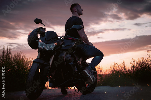 Young handsome male biker, is sitting on a motorbike and enjoying the sunset near the shore.