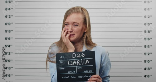 Mugshot of arrested young woman crying while holding sign for photo in police station. Millennial female person looking to camera while standing front of metric lineup wall.