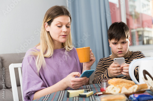 Portrait of young mother and son using phones during breakfast indoors