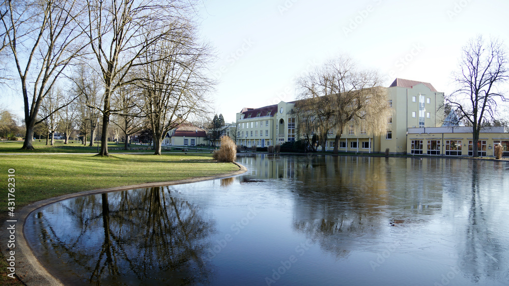 Obraz premium Jugendstil Kurhaus vor Seenfläche und kahlen Bäumen im Kurpark in Bad Hersfeld im Frühling mit Sonne ohne Menschen